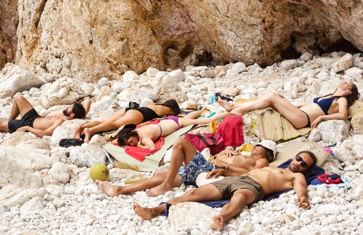 Un groupe de personnes, allongées sur une plage à cailloux inconfortables, dore au soleil.