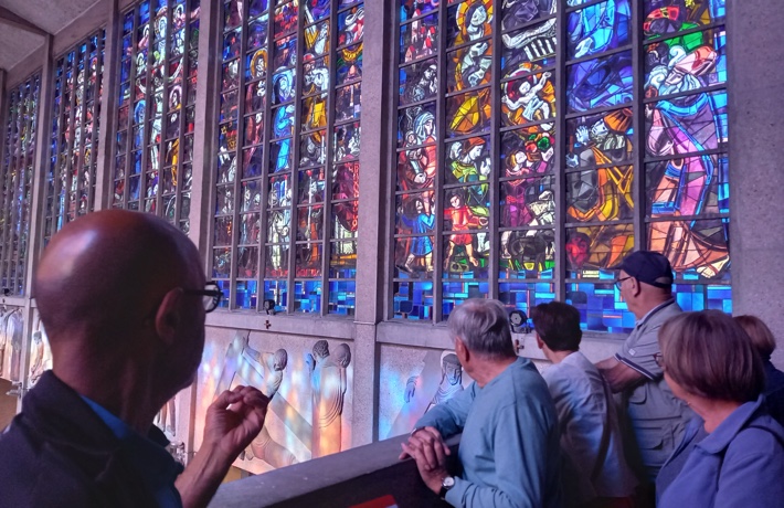 Une personne mène une visite autour de 5 autres, à l’intérieur de la basilique, face à des vitraux.