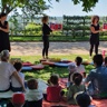 Trois femmes tiennent des livres dans leur main face à un groupe d’enfants avec leurs parents, sur la pelouse des jardins de l’évêché.