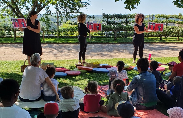 Trois femmes tiennent des livres dans leur main face à un groupe d’enfants avec leurs parents, sur la pelouse des jardins de l’évêché.