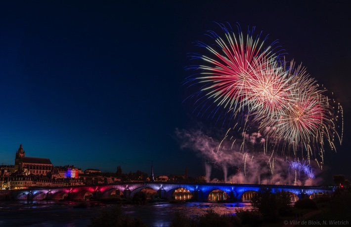 Feu d’artifice avec 3 grands panaches au-dessus du pont Jacques-Gabriel, chacun ayant du bleu, du blanc et du rouge. À l’arrière-plan, la cathédrale, puis l’Hôtel de Ville, ce dernier étant également illuminé en bleu-blanc-rouge.