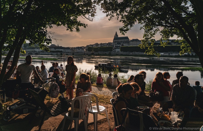 La foule est attablée sous les arbres du port de la Creusille pour dîner et discuter pendant un concert de la guinguette. La Loire et le cathédrale Saint-Louis sont à l’arrière-plan.