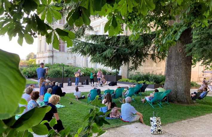 Des dizaines de personnes sont assises sur l’herbe ou les chaises des terrasses du château pendant qu’un groupe joue de la musique.