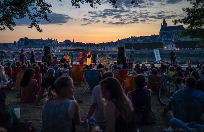 Un groupe en concert à la Creusille, avec et de nombreuses personnes devant lui jusqu’au premier plan, et la Loire et le centre-ville en arrière-plan tandis que le soleil couché fait crépuscule.