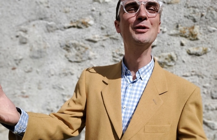 Un homme costumé et portant des lunettes sourit à l’objectif les bras écartés.